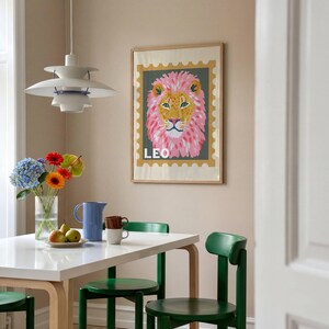 May include: A framed print featuring a lion with a pink mane and the word "LEO". The artwork is displayed above a white table with green chairs. A vase of flowers and a blue pitcher are on the table.