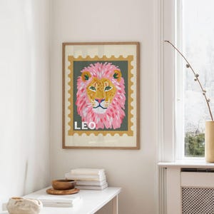 May include: A framed art print featuring a stylized lion's head with a pink mane and yellow face. The word "LEO" is printed in white at the bottom. The artwork is set against a green background with a postage stamp-like border.