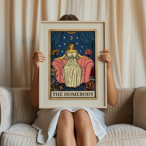 May include: A tarot card with a blue background and gold trim. The card depicts a woman sitting on a pink couch with a starry night sky behind her. The text "THE HOMEBODY" is printed in gold at the bottom of the card.