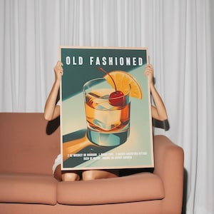 Op de afbeelding: Een ingelijste print met een illustratie van een "Old Fashioned" cocktail. Het kunstwerk toont een gedetailleerd glas met ijs, een kers en een sinaasappelschijf. Het recept staat onder de afbeelding. De achtergrond is teal en de lijst is licht hout.