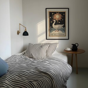 May include: A bedroom interior with a bed dressed in black and white striped bedding and pillows. A framed artwork with a goose illustration is on the wall. A small wooden side table holds a black jug and a white teacup. A brass and black wall lamp is mounted on the wall.