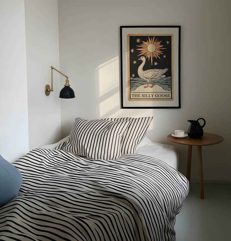 May include: A bedroom interior with a bed dressed in black and white striped bedding and pillows. A framed artwork with a goose illustration is on the wall. A small wooden side table holds a black jug and a white teacup. A brass and black wall lamp is mounted on the wall.