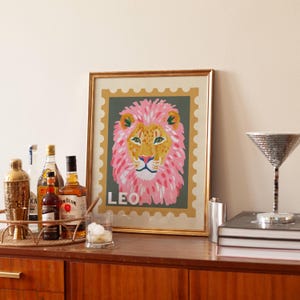 May include: A framed art print featuring a colorful lion illustration. The lion has a pink mane and is set against a green background with the word "LEO" at the bottom. The print is displayed on a wooden cabinet with bar accessories.