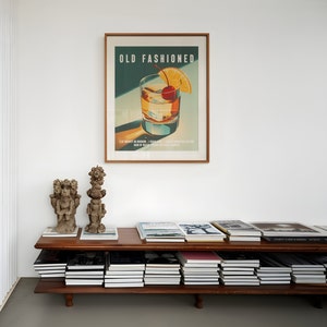 Op de afbeelding: Een ingelijste kunstprint getiteld "OLD FASHIONED" toont een cocktail met een kers en een sinaasappelschijfje. De print hangt boven een houten plank met boeken en twee kleine sculpturen. Het kleurenpalet omvat bruin, groen en oranje.