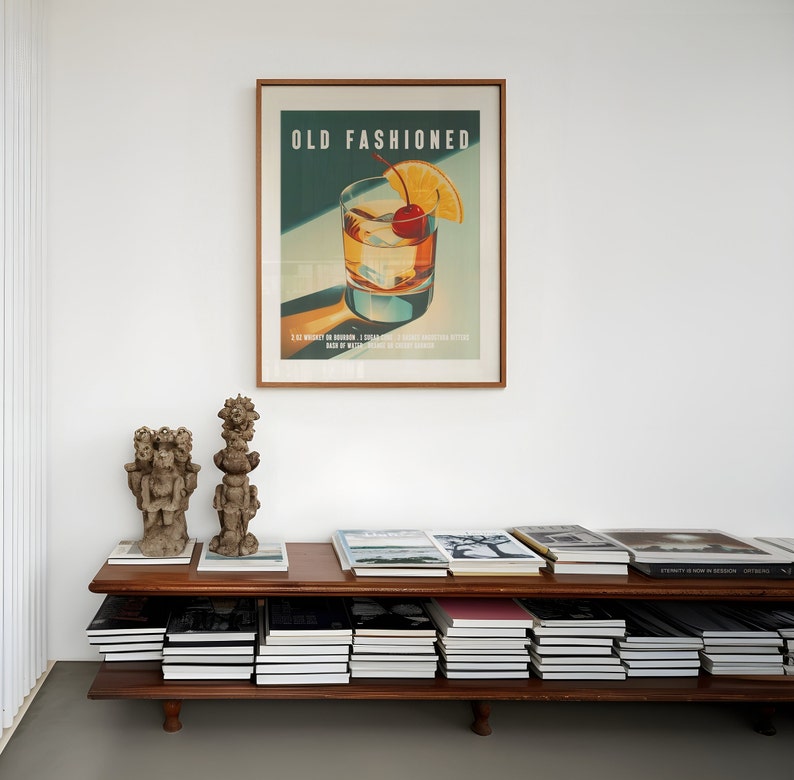 Op de afbeelding: Een ingelijste kunstprint getiteld "OLD FASHIONED" toont een cocktail met een kers en een sinaasappelschijfje. De print hangt boven een houten plank met boeken en twee kleine sculpturen. Het kleurenpalet omvat bruin, groen en oranje.