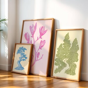 K&ouml;nnte beinhalten: Drei gerahmte Kunstdrucke, die an einer wei&szlig;en Wand lehnen. Die Drucke zeigen botanische Illustrationen: einen blauen Bonsai-Baum, rosa Magnolienbl&uuml;ten und eine gr&uuml;ne Waldszene. Die Rahmen sind aus hellem Holz.