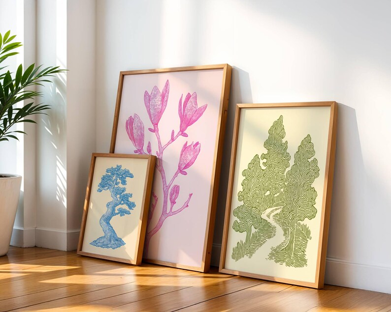 K&ouml;nnte beinhalten: Drei gerahmte Kunstdrucke, die an einer wei&szlig;en Wand lehnen. Die Drucke zeigen botanische Illustrationen: einen blauen Bonsai-Baum, rosa Magnolienbl&uuml;ten und eine gr&uuml;ne Waldszene. Die Rahmen sind aus hellem Holz.