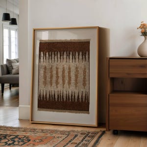 Könnte beinhalten: Ein gerahmtes Textilkunstwerk mit einer natürlichen Farbpalette. Das Kunstwerk zeigt ein gewebtes Design mit Braun- und Beigetönen, das in hellem Holz gerahmt ist. Das Stück wird in einem gut beleuchteten Raum vor einer weißen Wand präsentiert. Der Rahmen ist etwa 100 cm hoch.