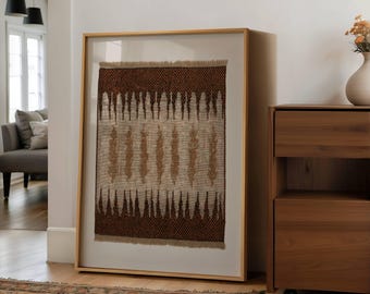 May include: A framed textile art piece with a natural, neutral color palette. The artwork features a woven design with brown and beige tones, framed in a light wood. The piece is displayed in a well-lit room, leaning against a white wall.