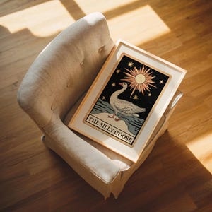 May include: A framed art print with a vintage illustration of a goose, with the text "THE SILLY GOOSE". The artwork features a sun, stars, and water elements against a dark background, creating a whimsical aesthetic.