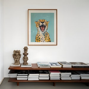 May include: A wooden shelf unit with two shelves, filled with books. On the top shelf, there are two decorative sculptures and a framed print of a cheetah with its mouth open. The print has a light blue background.