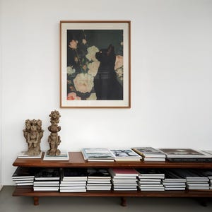 May include: A wooden shelf unit with two tiers, filled with stacks of books. Two small, decorative sculptures are on the top shelf, and a framed print of a black cat in a bouquet of flowers hangs on the wall above the shelf.