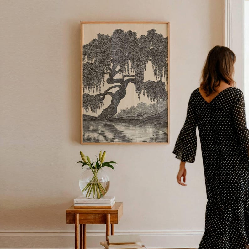 May include: A framed black and white print of a weeping willow tree over a body of water. The artwork is displayed on a light-colored wall. A small wooden side table with a glass vase of flowers is in the foreground.