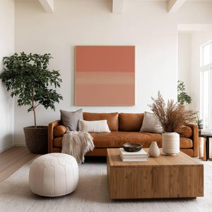 May include: A square abstract painting with horizontal bands of terracotta and peach tones hangs above a brown leather sofa. The room includes a wooden coffee table, a white round ottoman, a potted tree, and a vase with dried grasses.