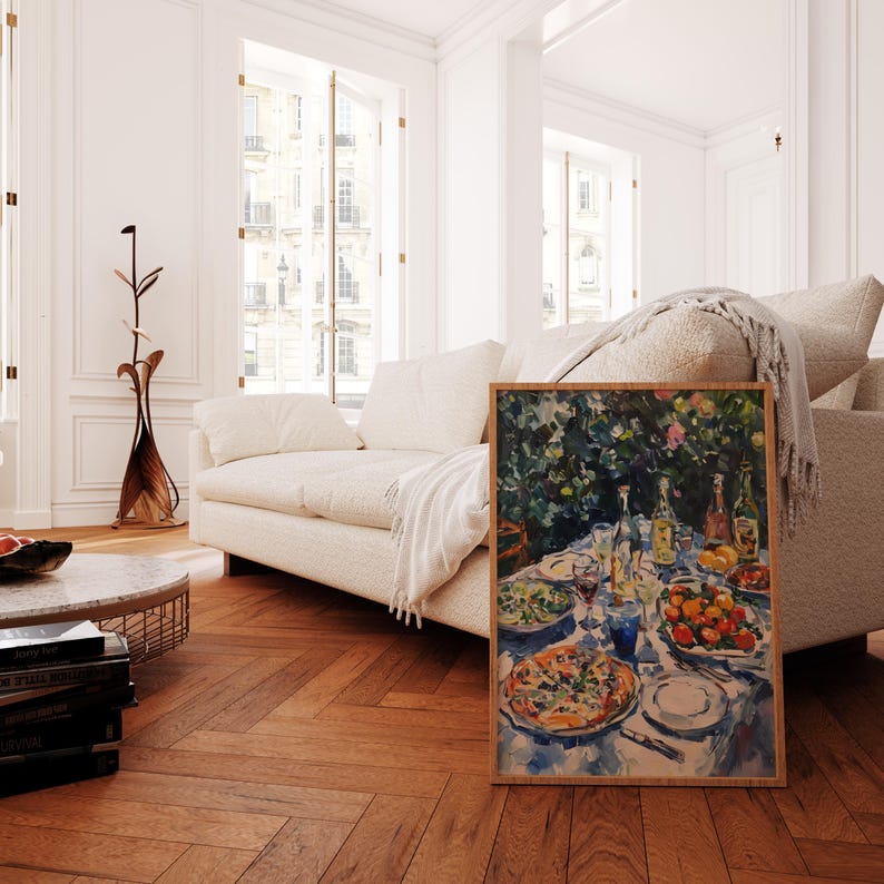 May include: A framed watercolor painting depicting a table setting with a pizza, salad, fruit, and wine glasses. The table is set with a blue tablecloth and surrounded by lush greenery.