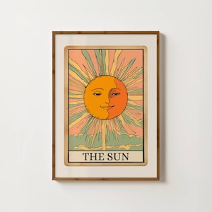 Può includere: Stampa artistica incorniciata di una carta dei tarocchi, raffigurante un sole sorridente con raggi arancioni, gialli e verdi. La carta è etichettata "THE SUN" in bianco su sfondo nero. La stampa è in una cornice di legno.