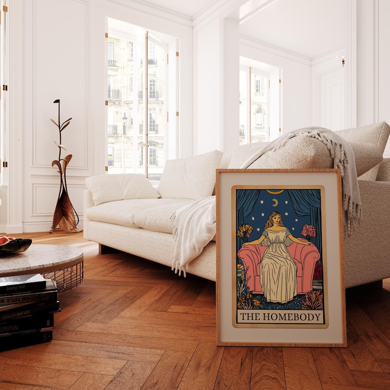 May include: A framed print of a tarot card illustration depicting a woman sitting on a pink couch with a crescent moon and stars in the background. The text "THE HOMEBODY" is printed at the bottom of the card.