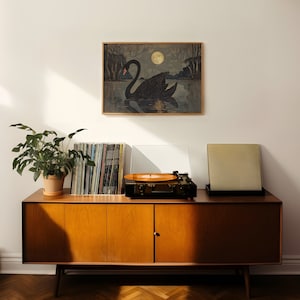 May include: A mid-century modern style wooden cabinet with three drawers and two doors. The cabinet is in a light wood tone and has a record player on top of it. There is a black and white framed print of a swan on the wall above the cabinet.