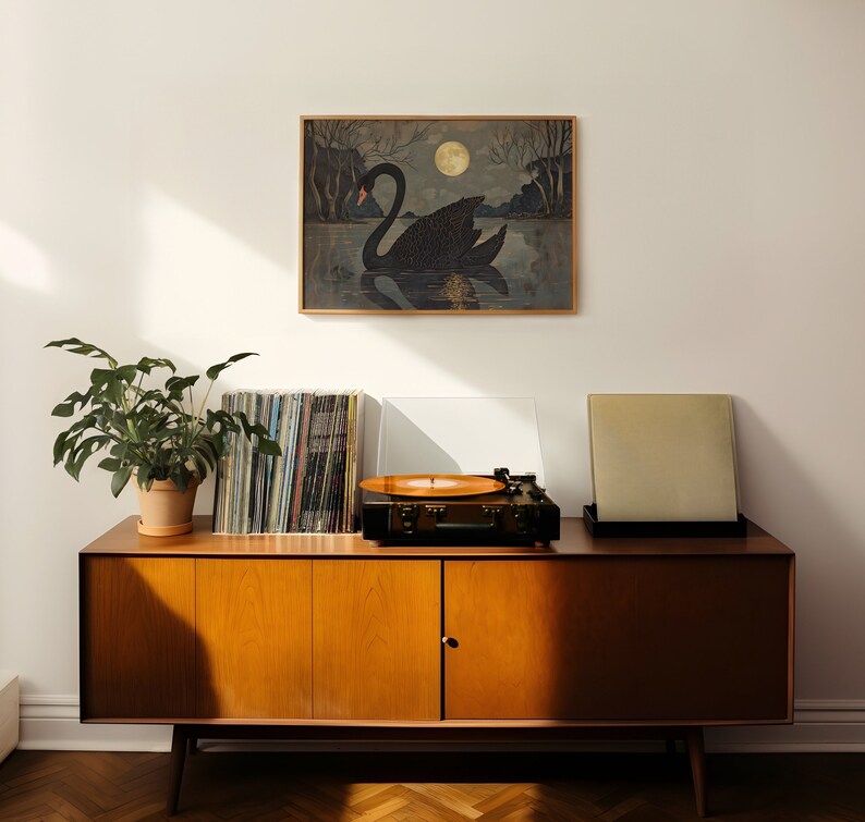 May include: A mid-century modern style wooden cabinet with three drawers and two doors. The cabinet is in a light wood tone and has a record player on top of it. There is a black and white framed print of a swan on the wall above the cabinet.