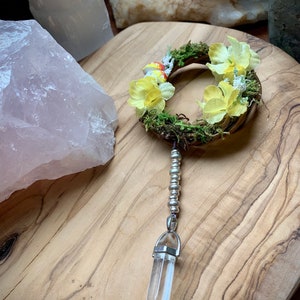 May include: A small, handmade wreath with yellow flowers and green moss, hanging from a silver chain with a clear quartz crystal pendant.