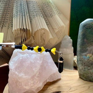 May include: A black and yellow beaded bracelet with a black crystal pendant. The bracelet is on a pink crystal rock and a book.
