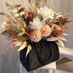 May include: A black handbag with a debossed monogram pattern serves as a vase for a large floral arrangement. The bouquet features white peonies, peach-colored pom-pom flowers, brown berry sprigs, dried grasses, and light green foliage stems.