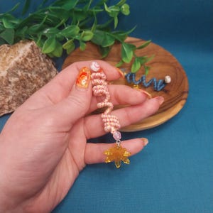 May include: A handmade, spiral-shaped earring with a yellow flower charm. The earring is made of pink and yellow thread, with a small pink bead and a white bead at the top. The flower charm is a translucent yellow color.