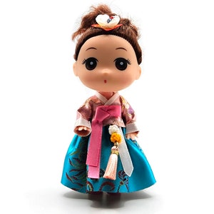 May include: A small doll wearing a traditional Korean hanbok dress. The dress is a vibrant blue with gold accents and a pink sash. The doll has brown hair and large, expressive eyes.
