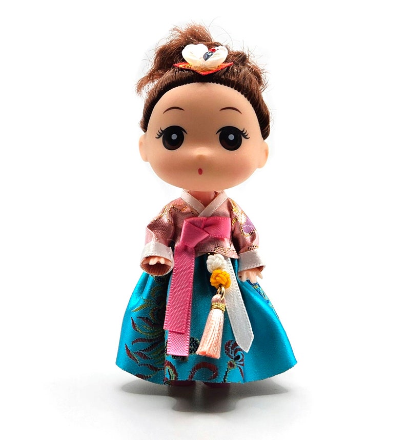 May include: A small doll wearing a traditional Korean hanbok dress. The dress is a vibrant blue with gold accents and a pink sash. The doll has brown hair and large, expressive eyes.