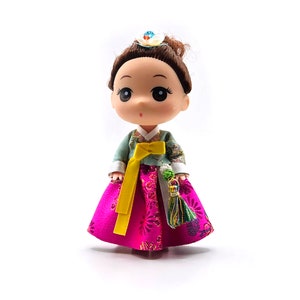 May include: A small doll wearing a traditional Korean hanbok dress. The dress is pink with floral patterns and a yellow sash. The doll has brown hair and large eyes.