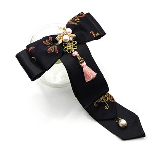 Pode incluir: La&ccedil;o de cetim preto com bordado dourado e vermelho, adornado com um amuleto de p&eacute;rola e ouro com uma borla rosa.