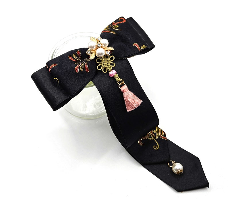 Pode incluir: La&ccedil;o de cetim preto com bordado dourado e vermelho, adornado com um amuleto de p&eacute;rola e ouro com uma borla rosa.