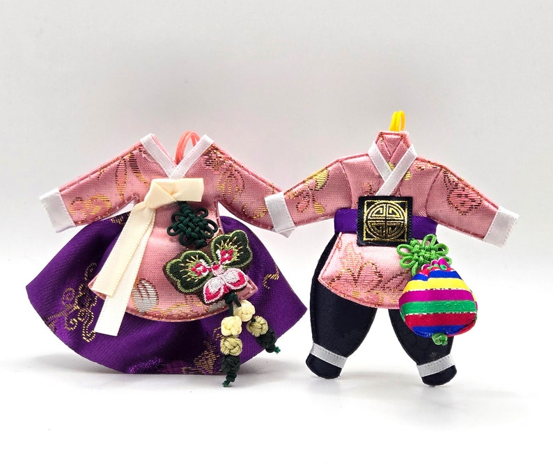 K&ouml;nnte beinhalten: Zwei traditionelle koreanische Miniaturkleidungsst&uuml;cke, ein rosa-violettes Kleid mit Schmetterlingsornament und ein rosa-schwarzes Outfit mit gestreiftem Beutel, beide mit komplexen Goldmustern, wei&szlig;en R&auml;ndern und dekorativen Stoffknoten.