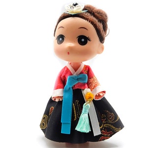 May include: A small doll wearing a traditional Korean hanbok dress. The dress is red and black with a blue sash and a white trim. The doll has brown hair and large eyes.