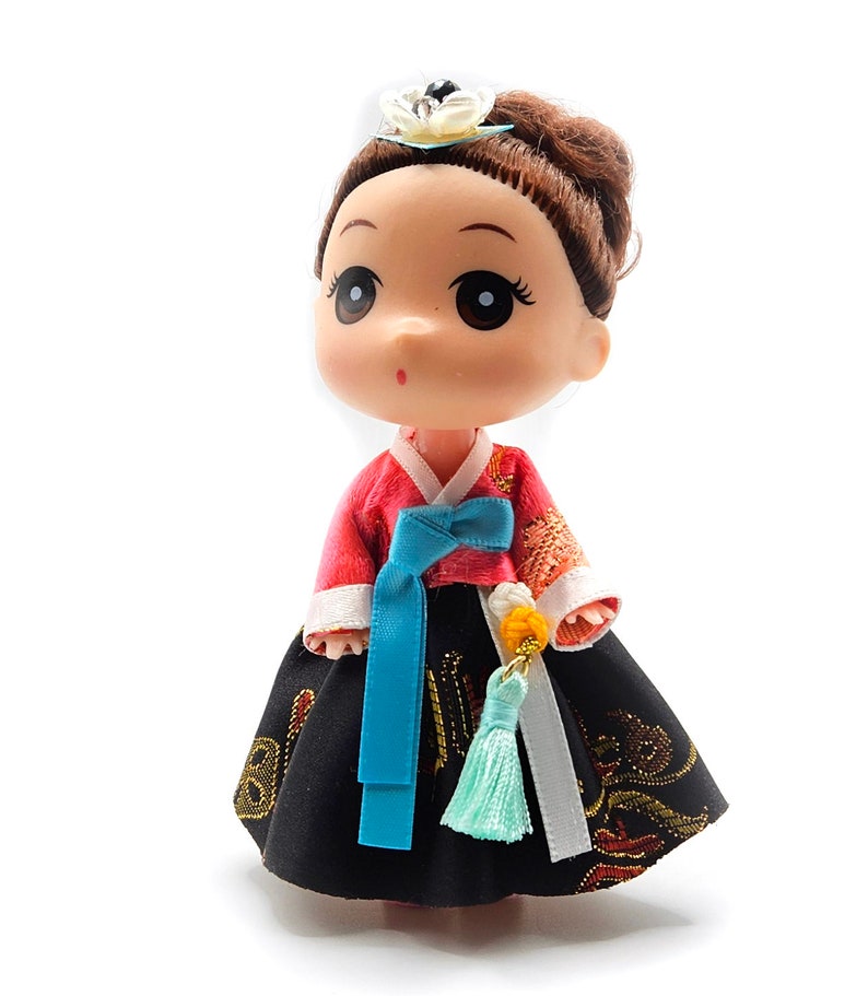 May include: A small doll wearing a traditional Korean hanbok dress. The dress is red and black with a blue sash and a white trim. The doll has brown hair and large eyes.