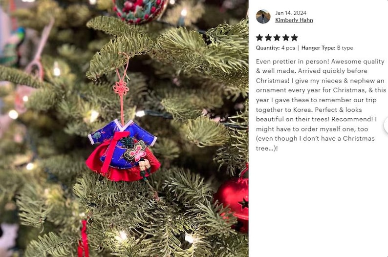K&ouml;nnte beinhalten: Ein Miniatur-Hanbok-Anh&auml;nger mit einem tiefblauen Oberteil mit wei&szlig;em Saum und einem leuchtend roten Rock. Das Kleidungsst&uuml;ck ist mit einem dekorativen Blumenaufn&auml;her und einer rosa Knotenschnur zum Aufh&auml;ngen an einem gr&uuml;nen Tannenzweig versehen.