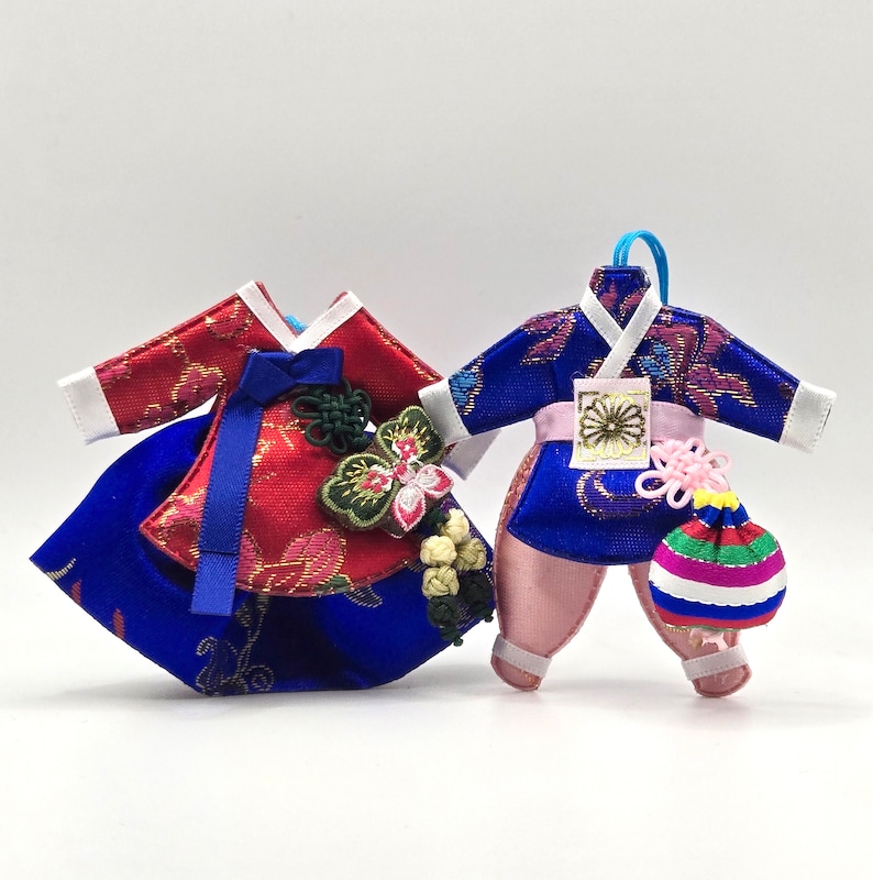 K&ouml;nnte beinhalten: Zwei Miniatur-Ornamente im traditionellen koreanischen Hanbok-Stil. Der linke Hanbok ist rot und blau mit Schmetterlingsverzierung und Knoten. Der rechte Hanbok ist blau und rosa mit goldenem Blumenemblem und einem kleinen gestreiften Beutel.