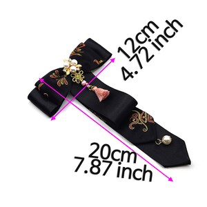 Pode incluir: Gravata borboleta preta com bordado dourado e vermelho, um detalhe de p&eacute;rola e borla, e medidas em cent&iacute;metros: 12 cm por 20 cm.