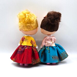 May include: Two dolls wearing traditional Korean hanbok dresses. The doll on the left is wearing a red and gold dress with a yellow jacket. The doll on the right is wearing a blue and gold dress with a pink jacket.