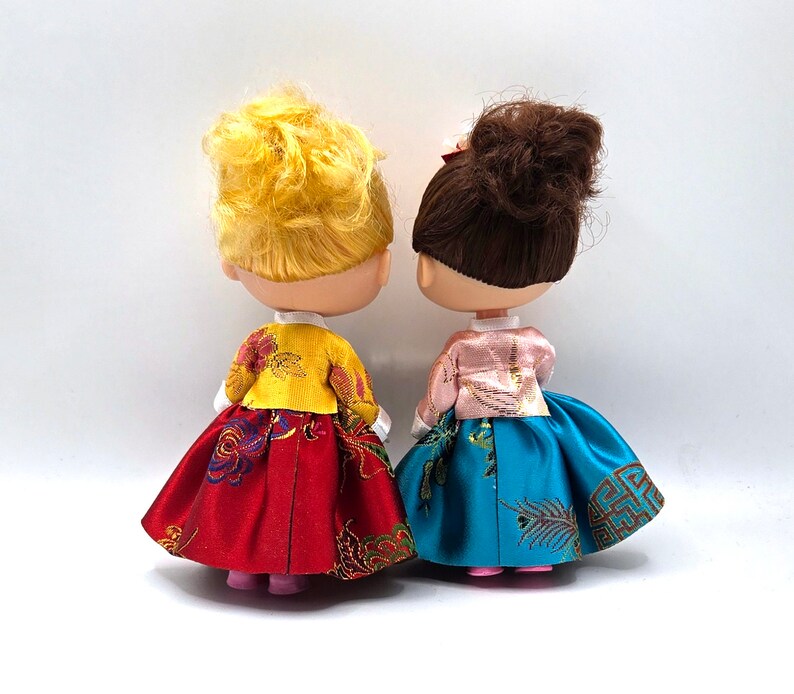 May include: Two dolls wearing traditional Korean hanbok dresses. The doll on the left is wearing a red and gold dress with a yellow jacket. The doll on the right is wearing a blue and gold dress with a pink jacket.