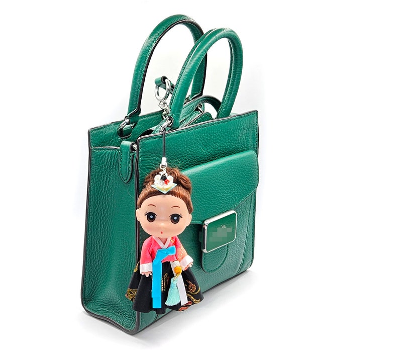 May include: A green leather handbag with a detachable strap and a small doll charm. The doll is wearing a traditional Korean dress.