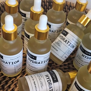 May include: A collection of small glass bottles with white labels and gold-colored caps. The labels read "HYDRATION" and "Hair Care." The bottles are filled with a light yellow liquid and are arranged in a woven basket.