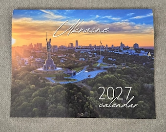 Op de afbeelding: Een kalender voor 2027 met een panoramische foto van het Moederland-monument in Kiev, Oekraïne, bij zonsondergang. De afbeelding toont het standbeeld, de skyline van de stad en groene bomen onder een oranje en blauwe lucht met de tekst Ukraine 2027 calendar.