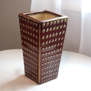 Puede incluir: Un jarrón cuadrado de cerámica con un esmalte rojo oscuro y detalles dorados. El jarrón tiene una superficie texturizada con pequeños puntos en relieve.