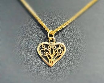 Peut inclure: Un collier pendentif doré avec un charm en forme de cœur et un motif ajouré. Le pendentif est suspendu à une chaîne fine. Le collier est présenté sur un fond sombre.