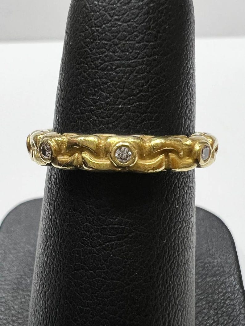 Puede incluir: Un anillo dorado con un dise&ntilde;o de eslabones de cadena. El anillo presenta peque&ntilde;as piedras preciosas redondas y transparentes engastadas a intervalos a lo largo de la banda. El anillo se muestra sobre un soporte negro sobre un fondo blanco.