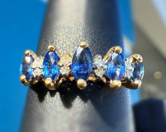 May include: A gold ring featuring marquise-cut blue gemstones and small clear stones. The ring is set on a dark surface, with a bright blue background. The gemstones are arranged in a repeating pattern.