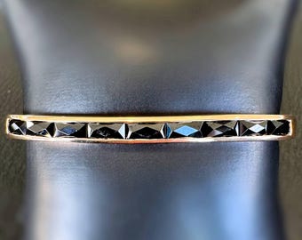 Peut inclure: Un bracelet doré orné d'une rangée de pierres noires taillées en forme géométrique. Le bracelet a une forme rectangulaire et est présenté sur une surface noire. Les pierres sont espacées uniformément et réfléchissent la lumière.