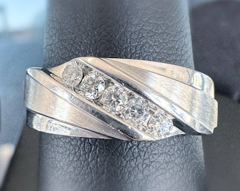 Peut inclure: Bague en argent avec une finition en métal brossé et une rangée de cinq pierres précieuses rondes et transparentes. La bague a un design moderne avec des rainures diagonales.