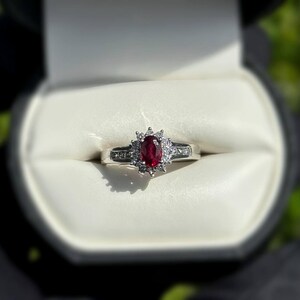 May include: A silver ring with a large oval ruby center stone, surrounded by smaller clear stones. The ring is set in a white box, with a blurred green background.